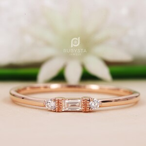 Peut inclure: Une bague en or rose ornée d'un diamant central taille baguette encadré par deux petits diamants taille ronde. Le sertissage présente des détails métalliques texturés de chaque côté de la pierre baguette. Le texte RUBYSTA JEWELS est visible.