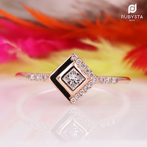 Peut inclure: Une bague en or rose avec un diamant central taille princesse serti dans un cadre carré, entouré d'un halo de petits diamants ronds. L'anneau est également orné de petits diamants ronds sur les côtés. Le texte RUBYSTA USA est visible.
