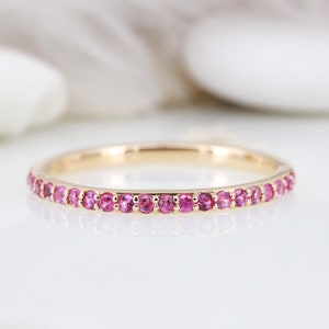 May include: A thin gold band ring featuring a continuous row of small, round, faceted pink gemstones set in a shared prong setting. The metal has a polished yellow gold finish, and the stones are uniform in size and vibrant pink in color.