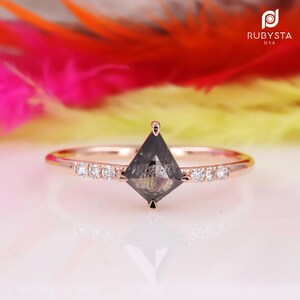 Peut inclure: Une bague de fiançailles en or rose avec un diamant noir de forme carrée et une rangée de diamants plus petits sur la bande.