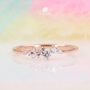 Peut inclure: Une bague en or rose ornée d'un groupe de sept pierres rondes transparentes taillées en brillant, disposées de manière asymétrique sur le dessus de l'anneau. Le logo RUBYSTA USA est visible au-dessus. Le métal présente une finition polie.