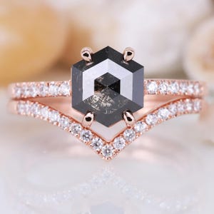 Peut inclure: Un ensemble de bagues de fiançailles en or rose avec une pierre centrale en diamant gris foncé de forme hexagonale. La bague est ornée de petits diamants clairs. La deuxième bague est en forme de V.