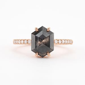 May include: A rose gold ring featuring a large, hexagonal, dark gray diamond as the centerpiece. The band is adorned with small, clear diamonds. The ring is set against a white background, highlighting the sparkle and design of the jewelry.