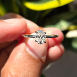 May include: A silver-toned ring featuring a single oval-cut clear gemstone set in a four-prong setting. The band is thin and polished, showcasing a reflective metallic finish that complements the faceted stone at the centre of the design.