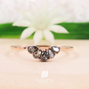 Peut inclure: Une bague en or rose présentant une disposition incurvée de cinq pierres en forme de poire, gris foncé et mouchetées. L'anneau est fin et poli, avec les pierres serties en arc symétrique au centre. Le texte RUBYSTA JEWELS est visible en dessous.