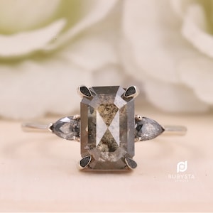 May include: A silver ring with a large, rectangular, gray, and black diamond. The diamond is set in a prong setting. The ring also has two smaller, pear-shaped diamonds on either side of the center stone.
