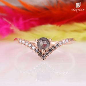 Peut inclure: Une bague en or rose avec un diamant gris central et des diamants plus petits sertis dans une bande en forme de V.