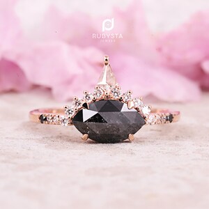 Peut inclure: Une bague en or rose avec une pierre centrale en diamant noir de forme marquise et un halo de diamants plus petits. La bague présente également un plus petit diamant de forme triangulaire sur le dessus de la bande.