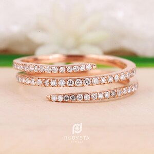 Peut inclure: Une bague en or rose au design à triple enroulement. L'anneau est incrusté de nombreuses petites pierres claires taillées en rond sur toute sa longueur. Le métal présente une finition métallique chaude et le texte RUBYSTA JEWELS est visible.