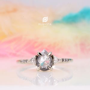 Peut inclure: Une bague en argent avec un diamant poivre et sel en forme de poire au centre, maintenu par quatre griffes. L'anneau est orné de deux petits diamants triangulaires de chaque côté et de petits diamants ronds incrustés sur la tige.