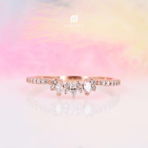 Peut inclure: Une délicate bague en or rose avec une rangée de petits diamants et un plus gros diamant de forme marquise au centre. La bague est posée sur un fond blanc.