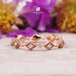 May include: A rose gold band ring featuring a repeating pattern of diamond-shaped settings, each containing a small round white gemstone. The metal surfaces are polished and textured with milgrain detailing. The logo RUBYSTA JEWELS is visible.