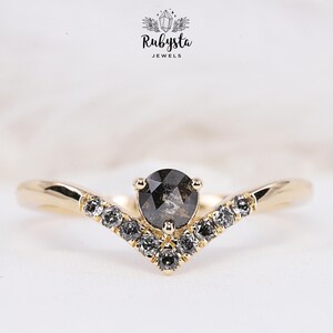 Peut inclure: Une bague en or avec un diamant noir central et une bande en V de diamants plus petits. La bague est posée sur un fond blanc.