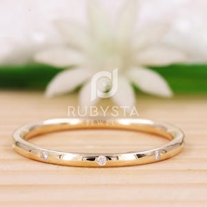 Peut inclure: Une fine bague en or avec de petites pierres rondes et transparentes serties à intervalles réguliers sur la surface extérieure. La bague a une finition polie et réfléchissante et est placée horizontalement. Le texte RUBYSTA JEWELS est visible.