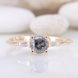 May include: A gold engagement ring featuring a round, grey diamond with two smaller, baguette-cut diamonds on either side.