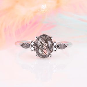 May include: A silver ring with a clear oval-shaped gemstone with black inclusions. The ring has two smaller clear gemstones with black inclusions on each side of the main stone.