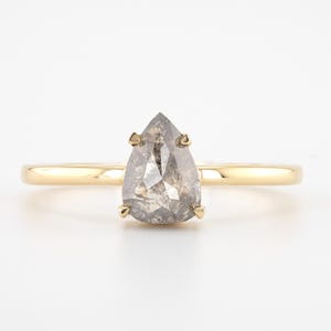 May include: A gold ring featuring a pear-shaped, gray diamond. The diamond is held in place by four prongs. The band is a simple, polished gold. The ring is set against a white background.