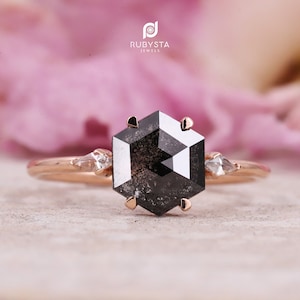 May include: A rose gold engagement ring with a hexagonal black diamond and two smaller accent diamonds. The ring is set against a pink floral background.