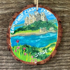 May include: A hand-painted wooden ornament featuring a coastal landscape. The artwork shows a castle, water, and the text "SOUTH WEST COAST PATH STAGE S". The ornament is round with a bark edge and a twine loop for hanging. The diameter is approximately 8 cm.