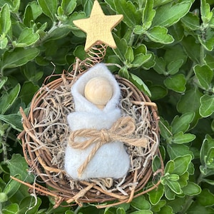 May include: A handcrafted Christmas ornament featuring a wooden baby swaddled in white felt, tied with twine, and nestled in a woven nest. A gold star tops the ornament, which is set against a backdrop of green foliage.