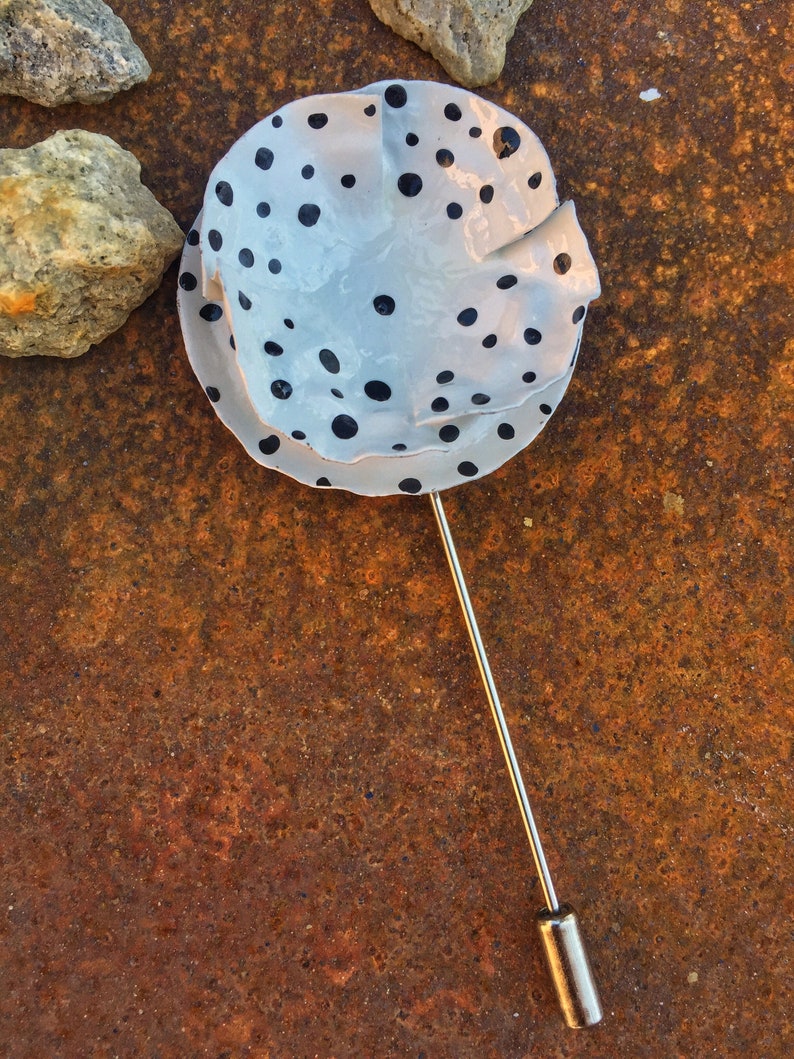 May include: A white ceramic pin with black polka dots. The pin is attached to a silver metal pin back.