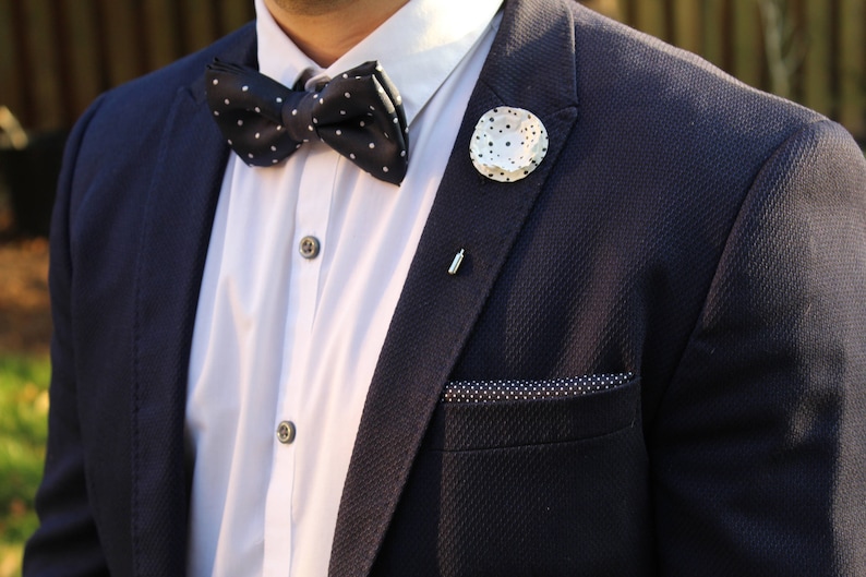 May include: A navy blue suit jacket with a white shirt, a black and white polka dot bow tie, and a white pin with a polka dot design.
