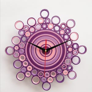 Orologio artigianale realizzato con la tecnica del quilling, composto da cerchi concentrici di carta nei toni del viola, glicine e del rosa. Il design ricorda un motivo floreale o una forma a raggiera, con un centro solido circondato da anelli di diverse dimensioni. Le lancette nere segnano l'ora. l'orologio è appeso ad un muro bianco