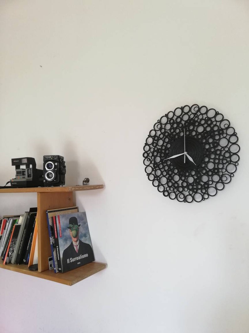 orologio è appeso di fianco ad una mensola con dei libri e delle macchine fotografiche