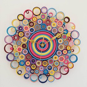 May include: A colorful clock made of paper rolls in shades of pink, blue, yellow, and red. The clock face is a concentric circle design with gold hands.