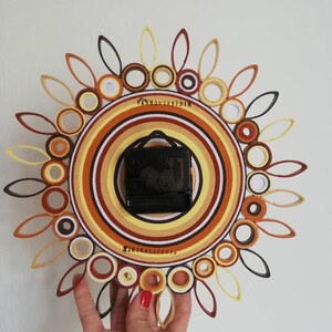 May include: A brown, orange, and white paper quilling clock with a black clock face. The clock is in the shape of a sun with a circle in the center. The clock is being held by a hand.