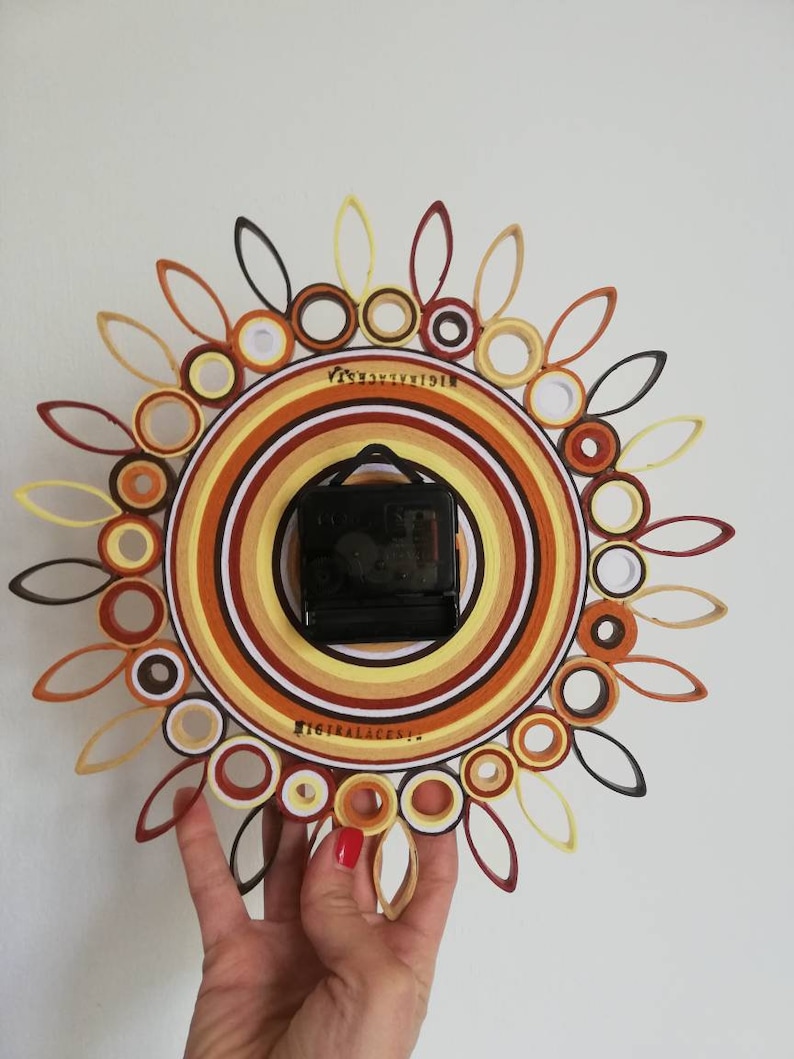 May include: A brown, orange, and white paper quilling clock with a black clock face. The clock is in the shape of a sun with a circle in the center. The clock is being held by a hand.