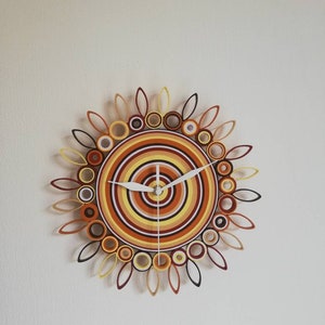 May include: A circular wall clock with a sunburst design made of rolled paper. The clock face is a concentric circle pattern in shades of orange, yellow, and brown. The clock hands are white.
