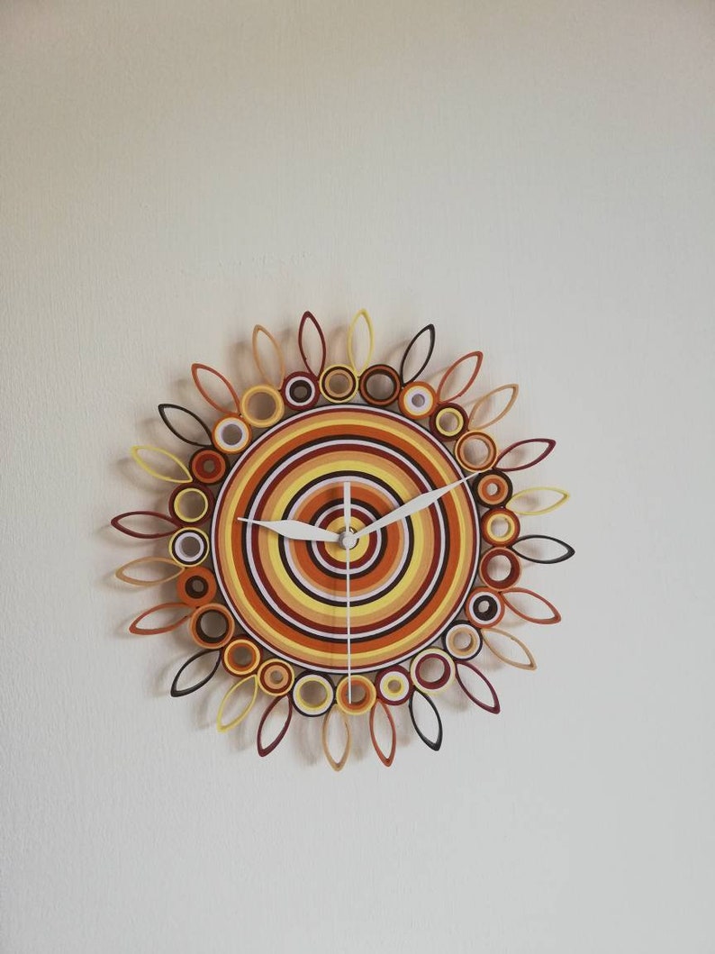 May include: A circular wall clock with a sunburst design made of rolled paper. The clock face is a concentric circle pattern in shades of orange, yellow, and brown. The clock hands are white.