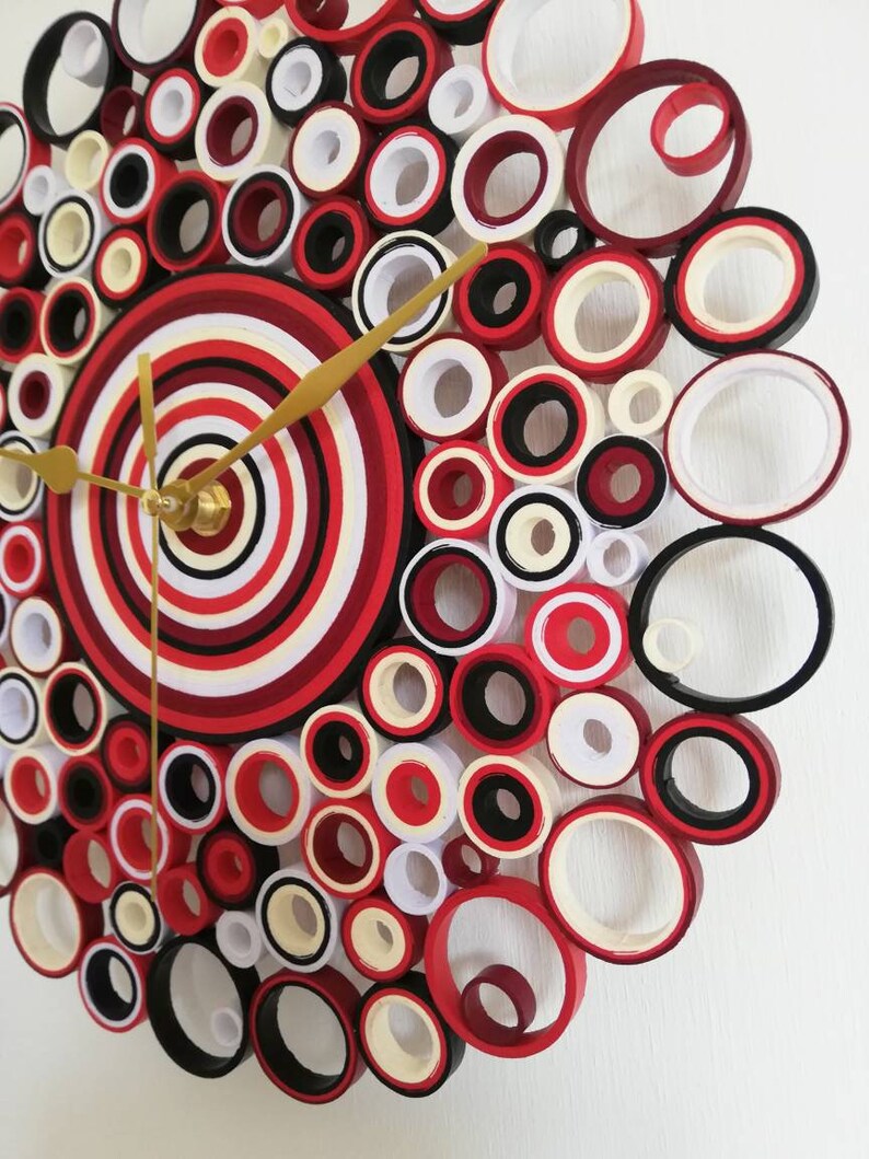 May include: A red, white, and black paper quilling clock with gold hands. The clock face is made of concentric circles of paper.