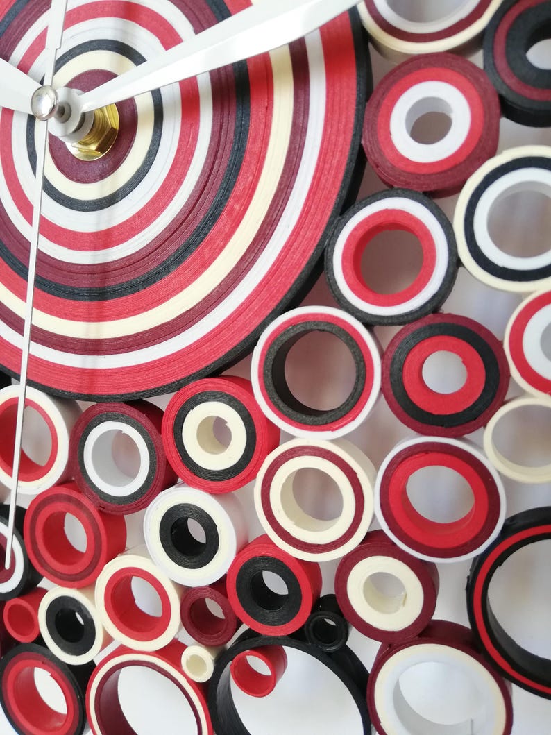 May include: A red, white, and black paper quilling clock with a circular design. The clock face is made of concentric circles of paper, and the hands are silver.