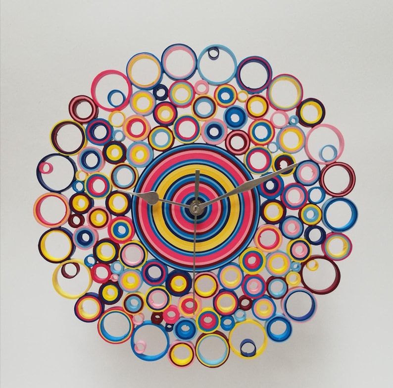 May include: A colorful clock made of paper circles in shades of pink, blue, yellow, and red. The clock face is a concentric circle design with a silver clock hand.