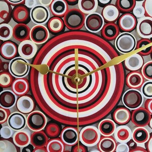 May include: A red, white, and black clock with a circular design. The clock face is made of concentric circles of paper, and the hands are gold. The clock is surrounded by a ring of smaller circles.