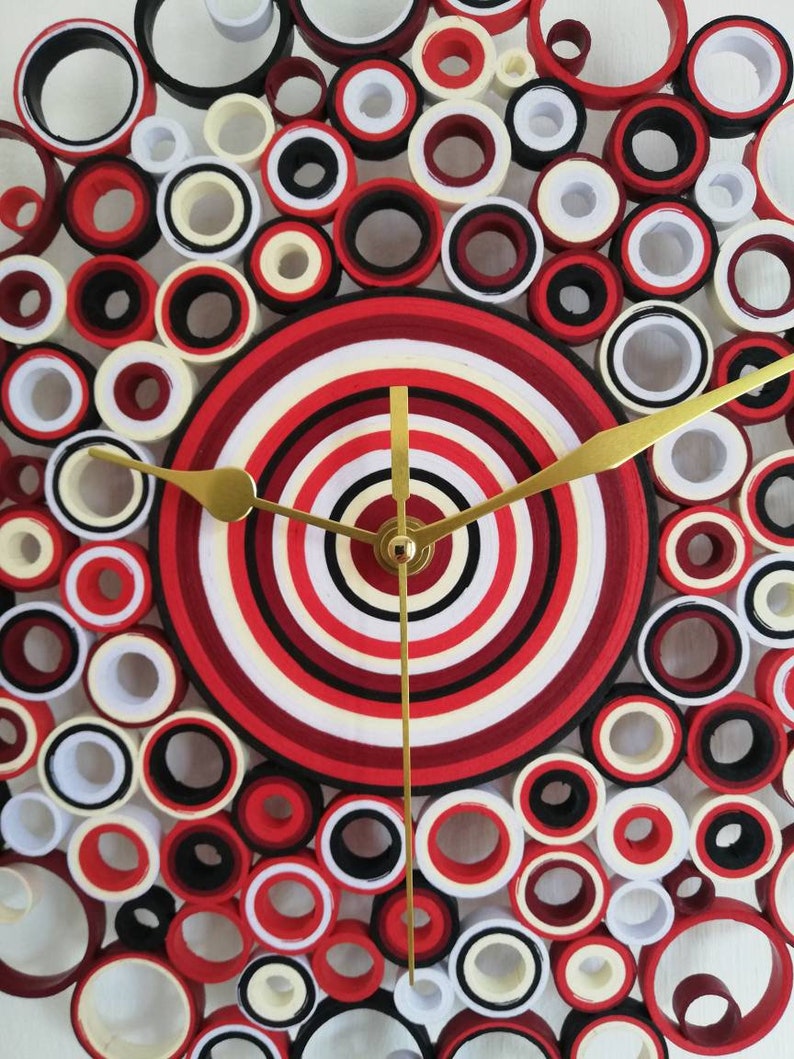 May include: A red, white, and black clock with a circular design. The clock face is made of concentric circles of paper, and the hands are gold. The clock is surrounded by a ring of smaller circles.