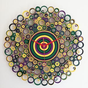 May include: A circular wall clock made of paper rolls, with a concentric circle design in green, yellow, and purple. The clock hands are red.