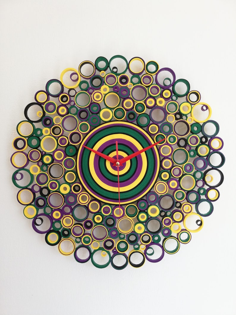May include: A circular wall clock made of paper rolls, with a concentric circle design in green, yellow, and purple. The clock hands are red.