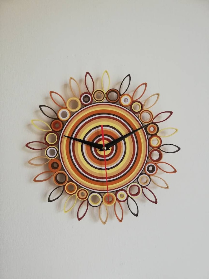 May include: A circular wall clock with a sunburst design. The clock face is made of concentric circles of brown, yellow, and white paper. The clock hands are black and red.