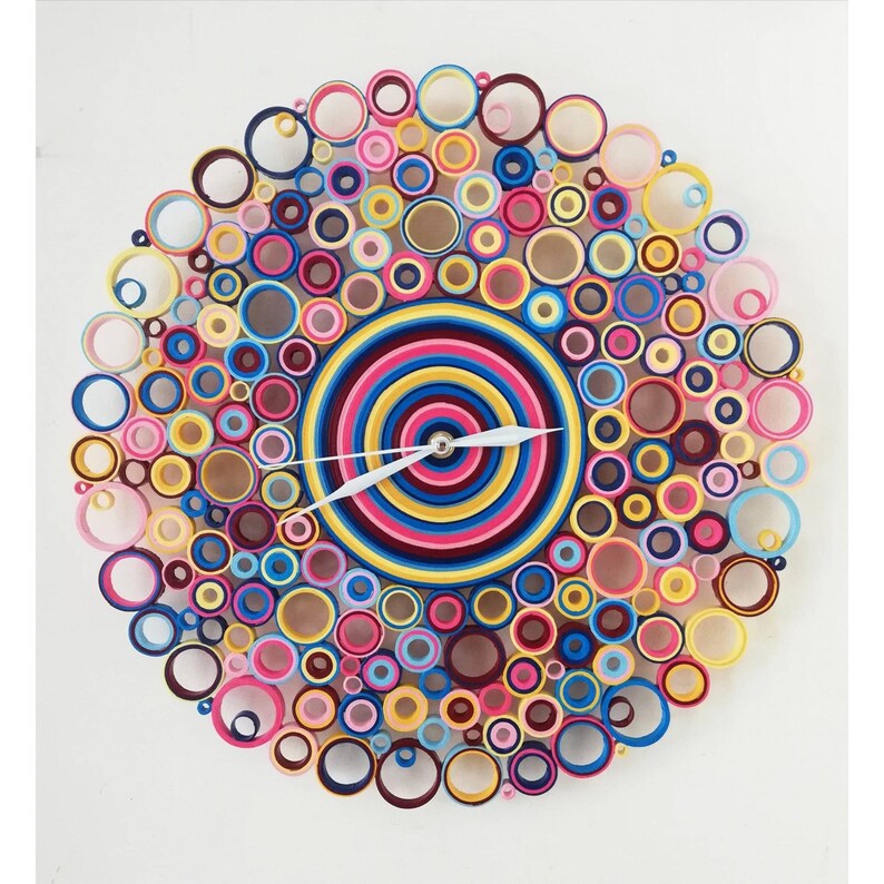 May include: A colorful clock made of paper rolls, with a white background. The clock face is a concentric circle design in shades of pink, blue, yellow, and red. The clock hands are white.