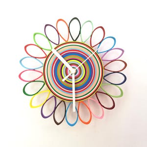 Op de afbeelding: Een kleurrijke wandklok met een bloemdesign. De klok heeft concentrische cirkels in verschillende kleuren en de buitenrand heeft bloemblad-vormige accenten in meerdere kleuren. Witte wijzers geven de tijd aan.
