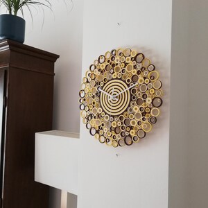 May include: A brown and yellow paper clock with a white face and hands. The clock is made of rolled paper circles and is hanging on a white wall.