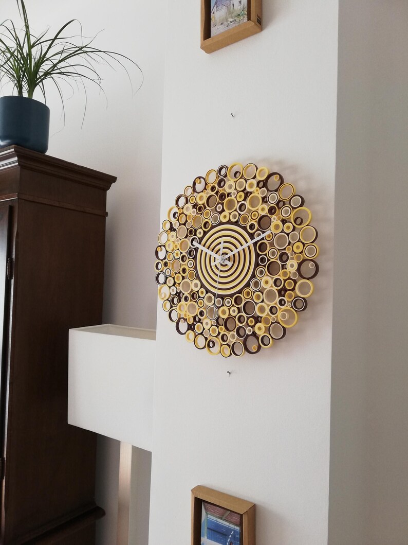 May include: A brown and yellow paper clock with a white face and hands. The clock is made of rolled paper circles and is hanging on a white wall.