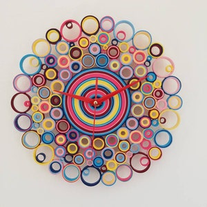 May include: A colorful clock made of paper circles in shades of pink, blue, yellow, red, and maroon. The clock face is a concentric circle design with a red hour hand and a black minute hand.