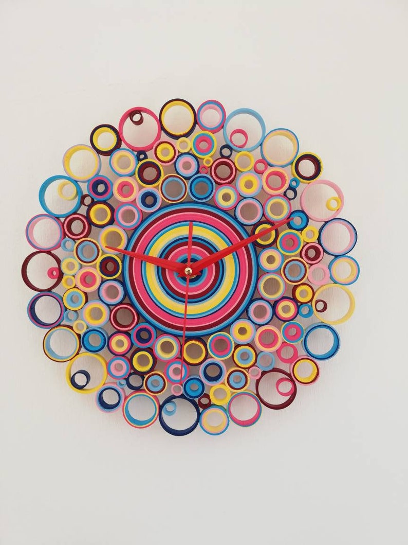 May include: A colorful clock made of paper circles in shades of pink, blue, yellow, red, and maroon. The clock face is a concentric circle design with a red hour hand and a black minute hand.