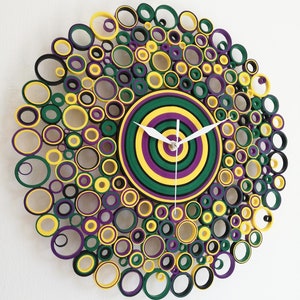 May include: A circular wall clock made of paper rolls, with a concentric circle design in shades of green, yellow, and purple. The clock face is a target-like design with white hands.