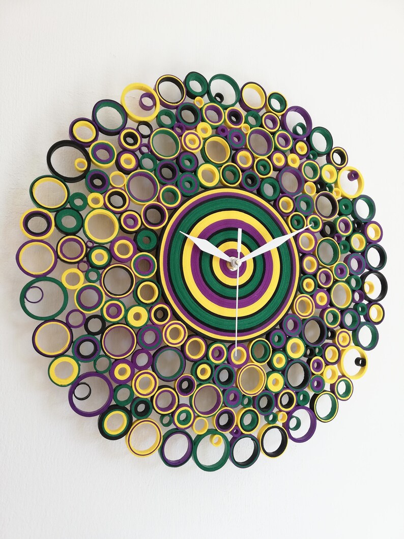 May include: A circular wall clock made of paper rolls, with a concentric circle design in shades of green, yellow, and purple. The clock face is a target-like design with white hands.