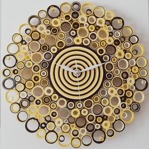May include: A yellow, brown, and white paper quilling clock with a circular design. The clock face is made of concentric circles of yellow and brown paper. The clock hands are white.